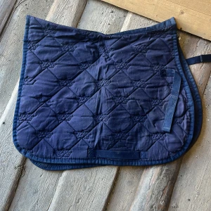Marinblått schabrak - Bilden visar en mörkblå, vadderad schabrak för häst, troligen av bomullsmaterial. Schabraket har quiltat mönster och remmar för fastsättning. Perfekt för dig som rider och vill ha en stilren och funktionell utrustning till din häst.