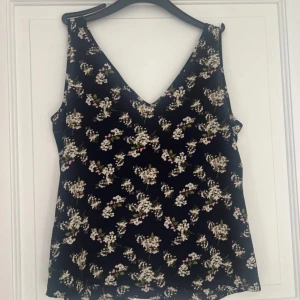 Blommig svart linnetopp med v-ringning - Snygg svart linnetopp med v-ringad hals och blommigt mönster i vitt och rosa. Toppen är ärmlös och har en avslappnad passform, perfekt för varma dagar. Materialet känns lätt och luftigt mot huden.
