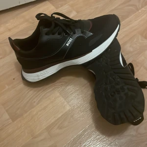 Svarta sneakers från BOSS - Snygga svarta sneakers från BOSS med vita sulor och diskreta röda detaljer. Skorna har snörning och en sportig design med både textil och syntetmaterial. Perfekta för dig som vill ha en clean och modern look med en touch av premiumkänsla.