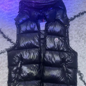 Moncler väst  - Säljer en svart, glansig dunväst från Moncler med huva och dragkedja framtill. Västen har två fickor med dragkedja och klassisk Moncler-logga på bröstet. Perfekt för lager-på-lager och riktigt snygg till streetwear. Står att den är 2xl men passar L