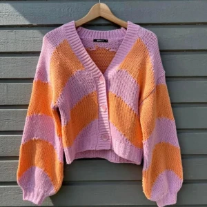 Randig kofta i lila och orange - Superfin stickad kofta med breda lila och orange ränder. Koftan är croppad med ballongärmar och stora knappar framtill. Perfekt för dig som vill sticka ut med färg och mönster. Snygg till både jeans och kjol!