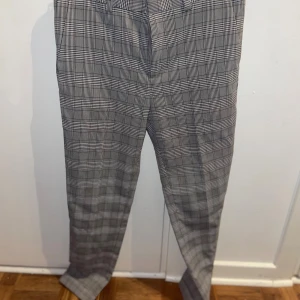 Grå rutiga kostymbyxor - Snygga grå kostymbyxor med klassiskt rutigt mönster. Byxorna har raka ben, bälteshällor och bakfickor med knappar. Perfekta för en clean och stilren look. Materialet känns mjukt och följsamt, passar dig som gillar dressad stil.