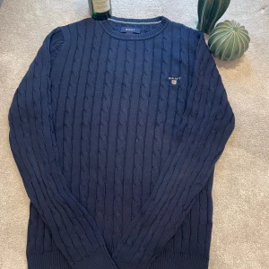 Mörkblå kabelstickad tröja från GANT - Kabelstickad mörkblå tröja från GANT med broderad logga på bröstet. Klassisk rund halsringning och ribbade muddar. Perfekt för dig som gillar preppy stil och vill ha något snyggt och tidlöst i garderoben.