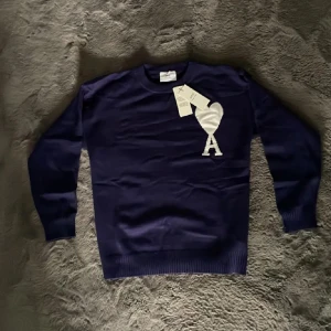 Ami Paris tröja  - Snygg mörkblå stickad tröja från Ami med stort vitt hjärta och A-logga på bröstet. Klassisk rund halsringning och ribbade muddar. Perfekt för dig som gillar stilrena detaljer och vill ha något som sticker ut lite extra.
