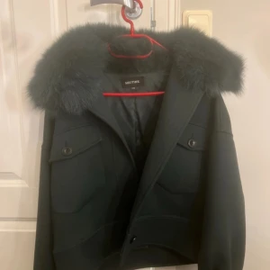 Svart jacka med päls från Meotine - Snygg svart jacka från Meotine med stor fluffig päls runt kragen. Jackan har två stora fickor framtill med knappar och en croppad passform. Perfekt för dig som vill sticka ut med en trendig look under kyliga dagar.