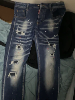 Blå distressed jeans från Dsquared2 - Säljer ett par blå jeans från Dsquared2 med slitna detaljer, hål och färgstänk. Jeansen har en rak passform och klassisk femficksdesign. Tillverkade i bomull och har en cool, edgy look som sticker ut. Pris kan diskuteras när det är snabb köp dom är äkta😁