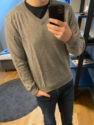 Grå stickad tröja från Morris - Snygg grå stickad tröja från Morris med V-ringning och diskret röd logga broderad på bröstet. Tröjan har långa ärmar och ribbade muddar vid ärmslut och nederkant. Perfekt att bära över en t-shirt eller skjorta. Den är lite nopprig, därav det låga priset. Storlek L, men lite bredare i passformen