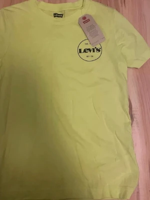 Gul Levi's t-shirt med logga - Fräsch gul t-shirt från Levi's med rund hals och svart logga tryckt på bröstet. Klassisk passform och korta ärmar, perfekt för en avslappnad stil. Tillverkad i mjuk bomull för skön känsla hela dagen.