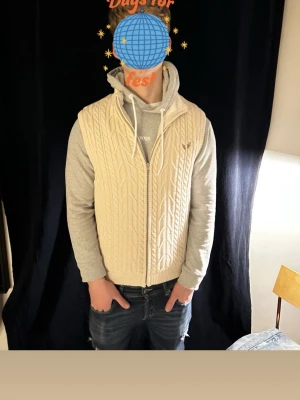Beige stickad väst med dragkedja - Snygg beige stickad väst med dragkedja framtill och klassiskt kabelstickat mönster. Perfekt att slänga över en hoodie för en avslappnad men trendig look. Västen har ribbade kanter och en mjuk känsla.
