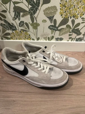 Vita Nike sneakers SB med svart swoosh - Snygga vita Nike sneakers med svart swoosh på sidan. Skorna har ovandel i mesh och mocka, vita snören och en platt sula. Perfekta för dig som gillar klassisk och sportig stil.