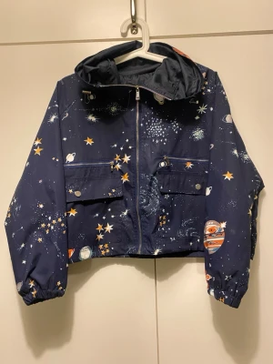 Blå vindjacka med rymdmönster - Japansk mörkblå vindjacka med rymdmönster av stjärnor, planeter och galaxer. Jackan har huva, dragkedja framtill, två stora fickor med knappar. Japansk stil. Kort och bred i modellen! 