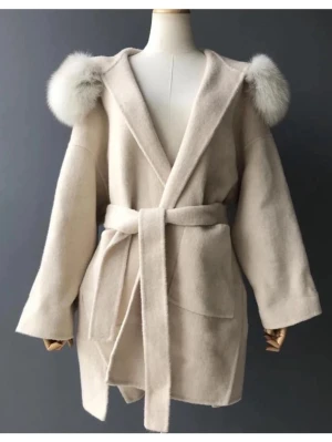 Beige kappa med äkta päls - Stilren beige kappa med stora fickor, knytskärp i midjan och fluffiga fuskpälsdetaljer på huvan. Kappan har en loose passform och är perfekt för kyligare dagar. Materialet är mjukt och ser ut att vara ullblandning.