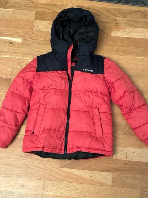 Röd och svart dunjacka från Everest - Snygg röd och svart dunjacka från Everest med huva och dragkedja framtill. Jackan har quiltad design, två sidofickor och reflekterande detalj bak. Perfekt för kalla dagar och har en sportig look.