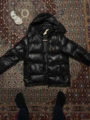 Svart pufferjacka från Polo Ralph Lauren - Snygg svart pufferjacka från Polo Ralph Lauren med huva och dragkedja. Jackan har det klassiska röda Polo-logot på bröstet och två sidofickor. Perfekt för kalla dagar och riktigt skön att bära. Passar dig som vill ha en stilren och varm jacka.