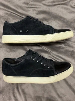 Svarta sneakers med mocka och lackdetalj - Säljer ett par svarta sneakers med ovandel i mocka och snygg lackad tå. Skorna har vita sulor, snörning och metallöljetter på sidan för extra ventilation. Perfekta för dig som gillar en stilren men ändå unik look.