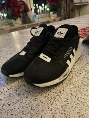 Svarta Adidas NMD sneakers - Snygga svarta Adidas NMD sneakers med vita detaljer och klassiska tre ränder på sidan. Skorna har snörning, rund tå och en bekväm, vit platt sula. Materialet är syntet och textil, perfekt för en sportig och modern look.