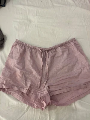 Ljusrosa pyjamasshorts - Säljer ett par ljusrosa pyjamasshorts med elastisk midja och dragsko framtill. Shortsen är tillverkade i ett tunt och mjukt tyg, perfekta för att chilla hemma, sova i eller ha på sommaren. De har en avslappnad passform och är superbekväma.