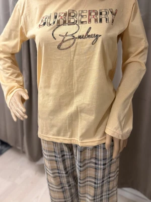 Beige Burberry pyjamas set - Mysig pyjamas från Burberry med långärmad beige tröja och logga framtill samt matchande byxor i klassiskt rutigt mönster i beige, svart och vitt. Perfekt för chillkvällar hemma. Mjukt material som känns skönt mot huden.