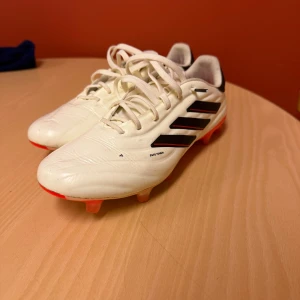 Vita Adidas fotbollsskor med svarta ränder - Säljer ett par vita Adidas fotbollsskor med klassiska svarta ränder på sidan och röda detaljer på sulan. Skorna har snörning och är tillverkade i syntetmaterial, perfekta för dig som vill ha stilrena och funktionella skor på planen.