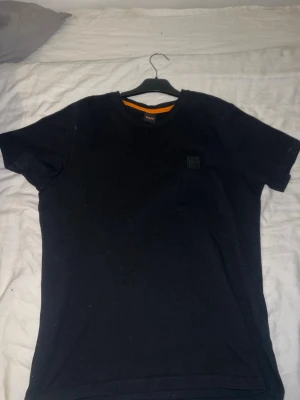 Svart t-shirt från Boss Orange - En stilren svart t-shirt från Boss Orange med rund halsringning och diskret logopatch på bröstet. Klassisk passform och tillverkad i mjuk bomull som känns skön mot huden. Perfekt till jeans eller shorts för en clean look.