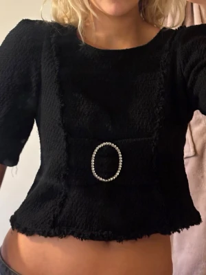 Svart croppad topp med strassdetalj - Trendig svart croppad topp från H&M med puffärmar och en stor oval strassdetalj framtill. Toppen har en lite ruffig textur och råa kanter. Jag är 180cm lång och bär vanligtvis S/M. Skicka gärna frågor eller prisförslag!