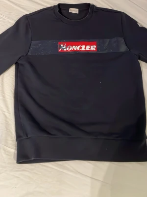 Svart sweatshirt från Moncler - Snygg svart sweatshirt från Moncler med tryckt logga i rött och vitt framtill. Tröjan har rund halsringning, ribbade muddar och ett Moncler-märke på ärmen. Storlek S