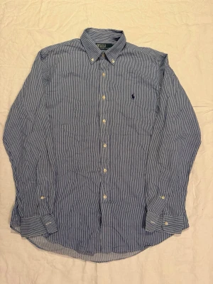 Blårandig skjorta från Polo Ralph Lauren - Klassisk blårandig skjorta från Polo Ralph Lauren med button-down krage och broderad logga på bröstet. Skjortan har långa ärmar, vita knappar och är tillverkad i mjuk bomull. Perfekt för en stilren och avslappnad look.