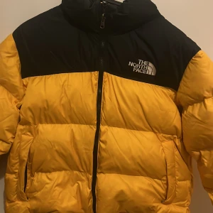 Gul och svart pufferjacka från The North Face prisförslag är välkomna  - Säljer en pufferjacka från The North Face i gul och svart. Jackan har broderad logga på bröstet, dragkedja framtill och två sidofickor. Den är quiltad och har hög krage som skyddar mot kyla. Perfekt för kalla dagar och riktigt snygg streetstil.