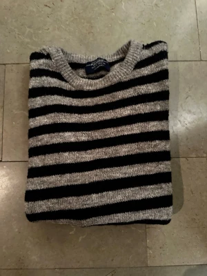 Randig stickad tröja  - Snygg stickad tröja från Polo Ralph Lauren med horisontella svarta och grå ränder. Tröjan har rund halsringning och långa ärmar. Tillverkad i mjukt garn som känns skönt mot huden. Perfekt för dig som gillar klassisk men avslappnad stil.