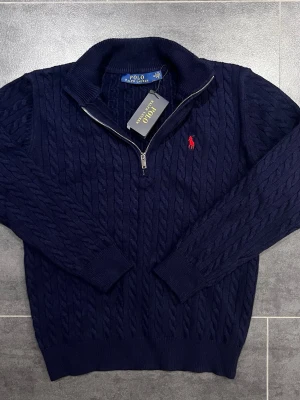 Mörkblå kabelstickad tröja Polo Ralph Lauren - Kabelstickad mörkblå tröja från Polo Ralph Lauren med halv dragkedja och hög krage. Tröjan har det klassiska röda Polo-logot broderat på bröstet och ribbade muddar vid ärmslut och nederkant. Perfekt för en stilren och avslappnad look.