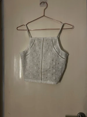 Vit spetsig linnetopp med blommönster - Söt vit linnetopp med tunna axelband och broderade blommor i spets. Toppen är croppad och har smockad rygg för extra komfort. Perfekt till sommaren och ger en fräsch och romantisk look.