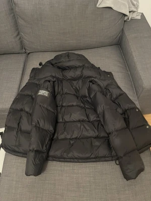 Svart pufferjacka från Burberry - Snygg svart pufferjacka från Burberry med hög krage och quiltad design. Jackan har Burberry London England-logga på ärmen och dragkedja framtill. Perfekt för kalla dagar och ger en clean, modern look.