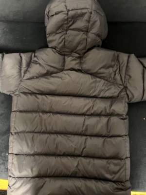 Svart dunjacka Peak Performance - Svart dunjacka från Peak Performance med huva och dragkedja framtill. Jackan har quiltad design och två sidofickor med dragkedja. Perfekt för kalla dagar och har en klassisk pufferlook.