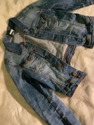 Klassisk blå jeansjacka - Säljer en klassisk blå jeansjacka med bruna knappar och två bröstfickor. Jackan har en tidlös design med långa ärmar och snygga söm-detaljer. Perfekt att slänga över en hoodie eller t-shirt för en chill look.