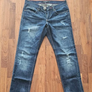 Philipp Plein Indian Cowboy Bootcut Jeans - W33  - Snygga blå bootcut jeans från Philipp Plein, modell Indian Cowboy. Jeansen har slitna detaljer, klassisk femficksdesign och cool döskalleknapp. Röd kontrastsöm och unika inslag på insidan ger extra edge. Perfekt för dig som gillar street och statement-plagg.
