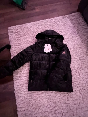 Svart Moncler jacka - Säljer en svart pufferjacka från Moncler med huva och coolt Moncler-märke på ärmen. Jackan har dragkedjor på fickorna och quiltad design som ger en schysst look. Insidan har en unik serietidningsetikett. Perfekt för dig som vill ha en varm och snygg jacka.