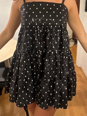 Svart kortklänning med paljetter - Supersöt svart kortklänning med vita prickar och glittriga paljetter. Klänningen har tunna axelband, volangig kjol och en loose passform som ger en skön och lekfull look. Perfekt för dig som vill sticka ut lite extra! Passar xs- m