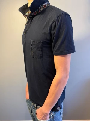 Dahlin skjorta - Mörkblå dahlin skjorta i bomull med korta ärmar och bröstficka. Kragen har ett färgglatt mönster på insidan som ger en unik touch. Perfekt för dig som vill ha en enkel men ändå speciell skjorta. Storlek M. Modellen på bilden är 184cm.