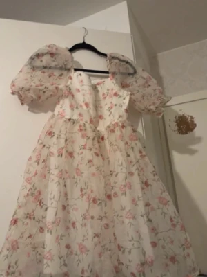 Blommig långklänning med puffärm - Romantisk långklänning i vitt med rosa blommönster och puffiga, transparenta ärmar. Klänningen har en markerad midja och voluminös kjol, perfekt för sommarens alla tillfällen. Tillverkad i ett lätt, luftigt material som ger en drömmig känsla.