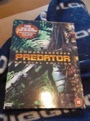 Predator Special Edition DVD - 2-disc specialutgåva av filmen Predator med Arnold Schwarzenegger. Omslaget är svart och grönt med futuristiskt motiv och tydlig gul text. DVD-fodralet har åldersmärkning 18 år och innehåller extramaterial. Perfekt för dig som gillar action och klassiska filmer.