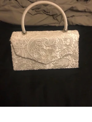Vit spetskuvertväska med handtag från KOKO - Säljer en elegant vit kuvertväska med spetsdetaljer och ett stadigt handtag. Väskan har ett vackert blommigt spetsmönster som ger en lyxig känsla. Perfekt för dig som vill ha något extra till din outfit. Väskan är från koko men saknar en axelrem. Meddela mig vid frågor, priset kan diskuteras 💕