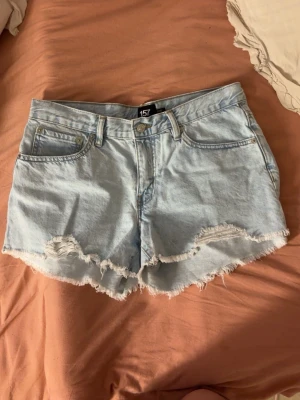 Ljusblå jeansshorts från lager 157 - Säljer ett par ljusblå jeansshorts från lager 157 med råa, fransiga kanter och klassisk femficksdesign. Shortsen har bälteshällor och knappgylf. Perfekta för varma dagar och ger en avslappnad vibe.