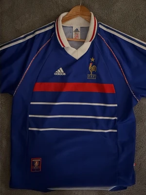 Blå Adidas Frankrike fotbollströja - Klassisk blå fotbollströja från Adidas med Frankrikes landslagslogga (FFF) och vita samt röda detaljer. Tröjan har korta ärmar, vit krage med röd och blå rand, samt vita ränder på axlarna. Tillverkad i polyester och perfekt för fotboll eller samling.
