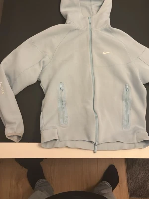 Ljusblå Nike NOCTA hoodie - Snygg ljusblå hoodie från Nike NOCTA-kollektionen med dragkedja framtill, två fickor med dragkedja och broderad Nike-logga på bröstet. Huva och detaljerade sömmar ger en sportig vibe. NOCTA-tryck på ärmen och diskreta vita initialer på ryggen.