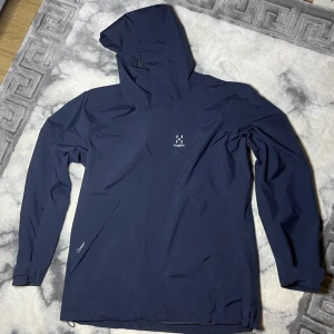 Mörkblå regnjacka från Haglöfs - Snygg oanvänd mörkblå regnjacka från Haglöfs med huva och lång ärm. Jackan har minimalistisk design, diskret logga på bröstet och dragkedjeförsedd ficka. Perfekt för regniga dagar och outdoor-äventyr. Vid frågor hör av dig!