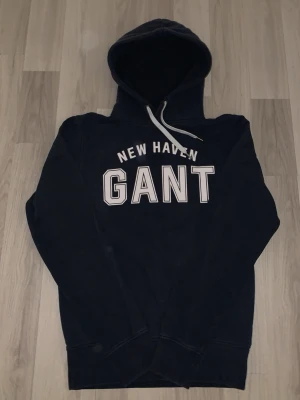 Mörkblå hoodie från GANT - Mörkblå hoodie från GANT med vit text 'NEW HAVEN GANT' på bröstet. Klassisk huva med vita dragsnören och ribbade muddar vid ärmslut och nederkant. Tillverkad i mjukt bomullsmaterial som känns skönt mot huden. Perfekt för en avslappnad och stilren look.