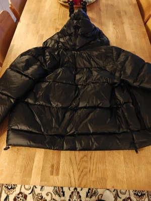 Svart dunjacka med röd mönstrad insida - Snygg svart dunjacka med huva och dragkedja. Insidan har ett unikt rött mönster med svarta detaljer som ger extra edge. Jackan är quiltad och har en oversized passform, perfekt för kalla dagar.