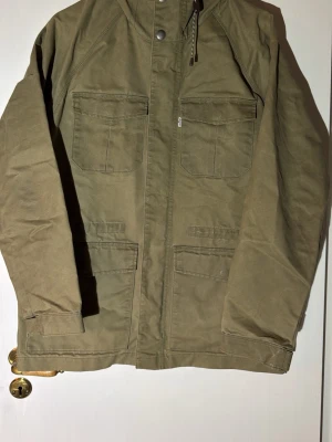 Olivgrön jacka från Levi's med fickor - Snygg olivgrön jacka från Levi's med fyra stora fickor framtill, dragkedja och tryckknappar. Jackan har huva med snörning och är tillverkad i slitstark bomull. Perfekt för dig som gillar en enkel men stilren look.