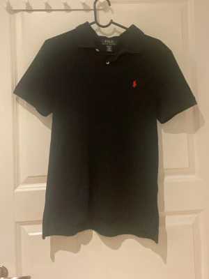 Svart pikétröja från Ralph Lauren - Klassisk svart pikétröja från Polo Ralph Lauren med röd broderad logga på bröstet. Tröjan har korta ärmar, krage och knappar vid halsen. Perfekt till en clean och stilren look.