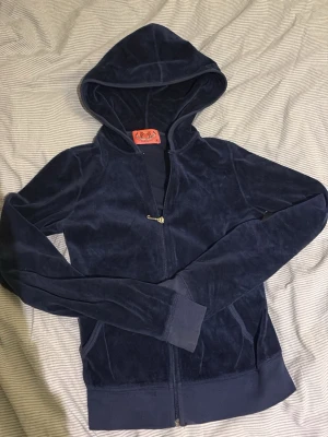 Juicy Couture - Snygg mörkblå hoodie från Juicy Couture i mjuk velour. Klassisk modell med dragkedja framtill, huva och fickor på sidorna. Perfekt för en avslappnad och trendig look. Ikonisk logga på insidan av kragen.